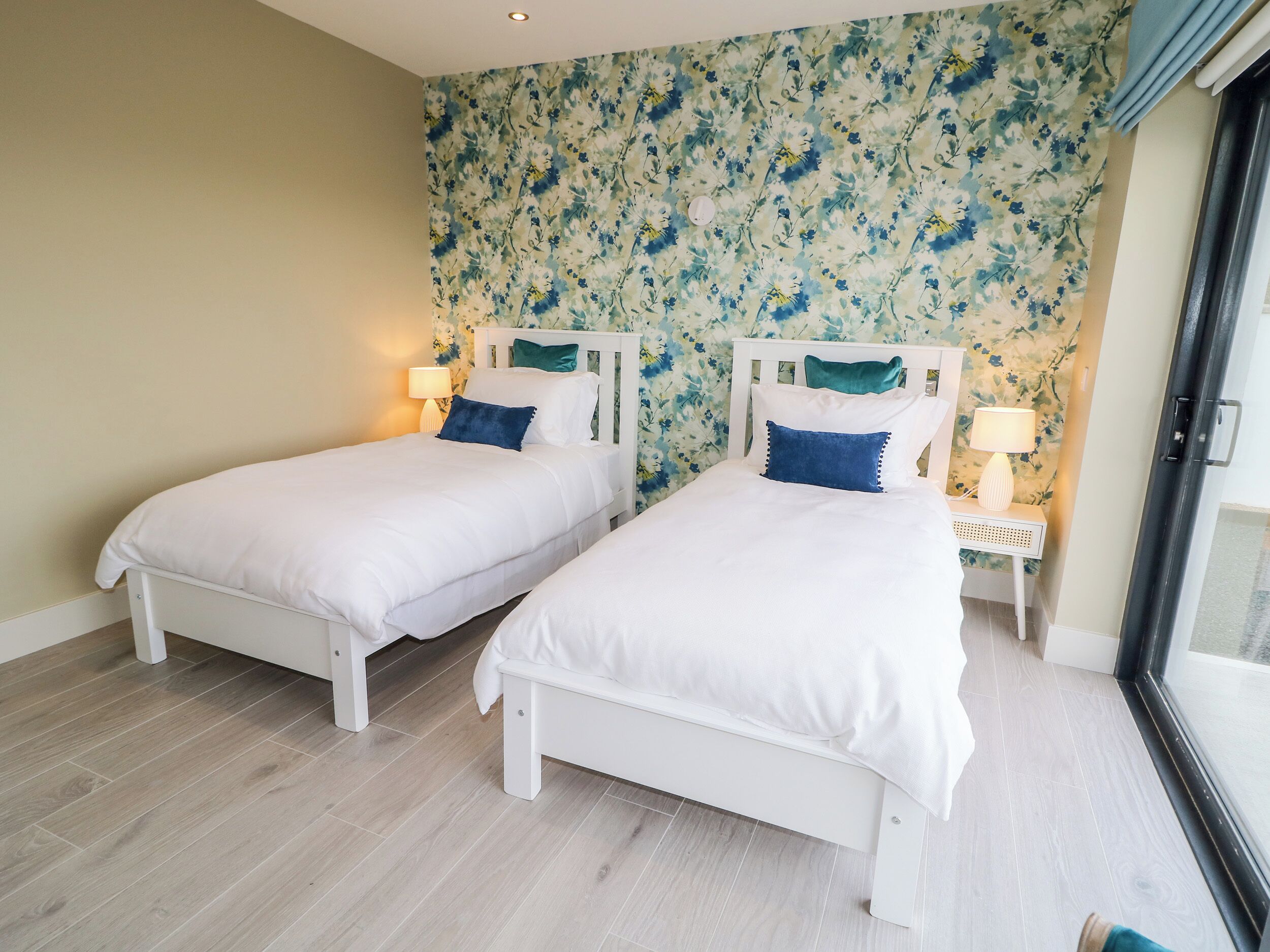 Ein Schlafzimmer mit zwei weißen Betten, blumigem Wandpapier und einem Fenster mit Blick.