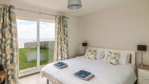 A bedroom with a large bed, two bedside tables, a window with curtains, and a view of the outdoors. - Beach House (Courtown)
