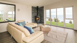 A modern living room with a beige sofa, a fireplace, and a large window overlooking the sea. - Beach House (Courtown)