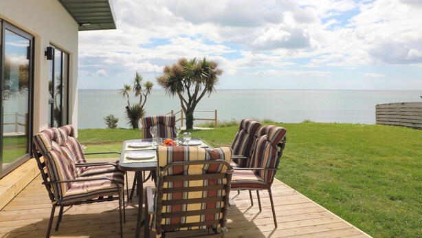 An outdoor patio with a table set for four, surrounded by striped chairs, overlooking a body of water. - Beach House (Courtown)