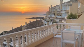 Un balcon agrémenté de meubles en osier, d’une table en verre et de chaises. La vue offre un panorama sur le coucher de soleil sur la mer et sur une ville côtière.