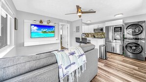 A modern living room with a grey sofa, a mounted TV displaying a beach scene, a ceiling fan, and a kitchen area with stainless steel appliances. - Victoria Park Gem! 1 Mile To Beach & Las Olas! (Fort Lauderdale)