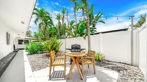 A patio with a round table and two chairs, surrounded by a white fence, with a grill and plants in the background. - Victoria Park Gem! 1 Mile To Beach & Las Olas! (Fort Lauderdale)