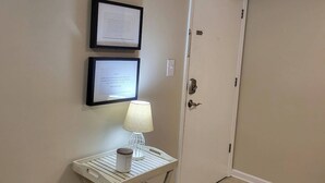 Un pasillo con una puerta blanca, un certificado en un marco en la pared y una pequeña mesa con una lámpara.
