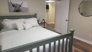 Un dormitorio con cama, almohadas, una mesita de noche, lámpara, un cuadro en la pared, una puerta y un espejo.