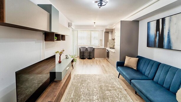 A modern living room with a blue sofa, a coffee table, and a TV. There is a kitchen in the background with a dining area.