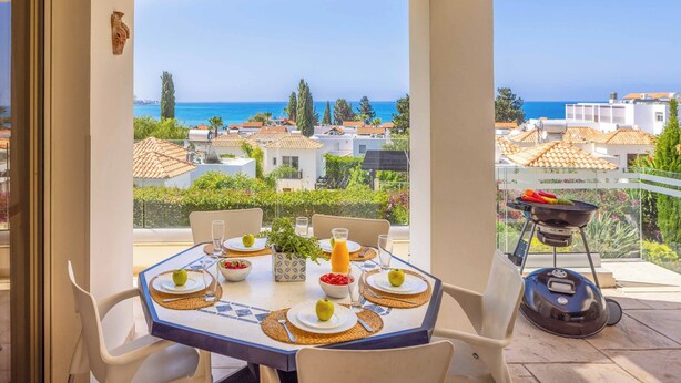 Una mesa de comedor preparada con platos, vasos y una parrilla en un balcón con vista al mar y edificios.