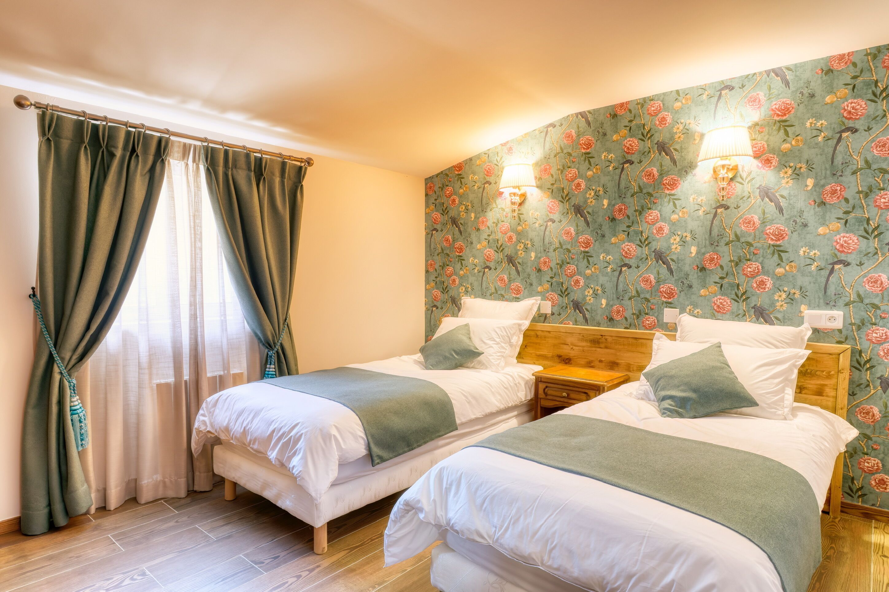 A room with two beds, wooden headboards, a floral wallpaper, and a window with curtains.