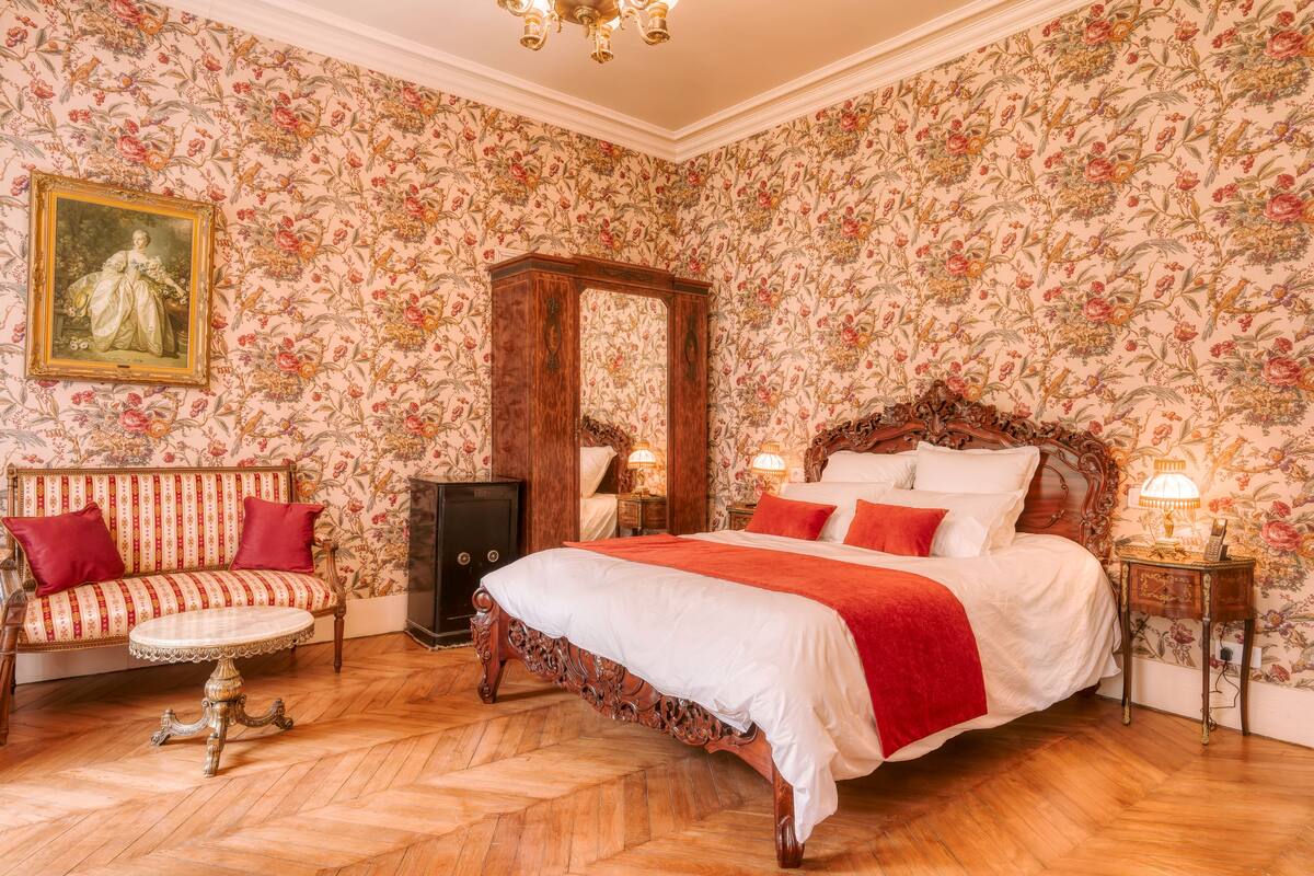 Une chambre à coucher classique, avec un grand lit, une table de chevet en bois, une table ronde et un canapé agrémenté de coussins rouges.