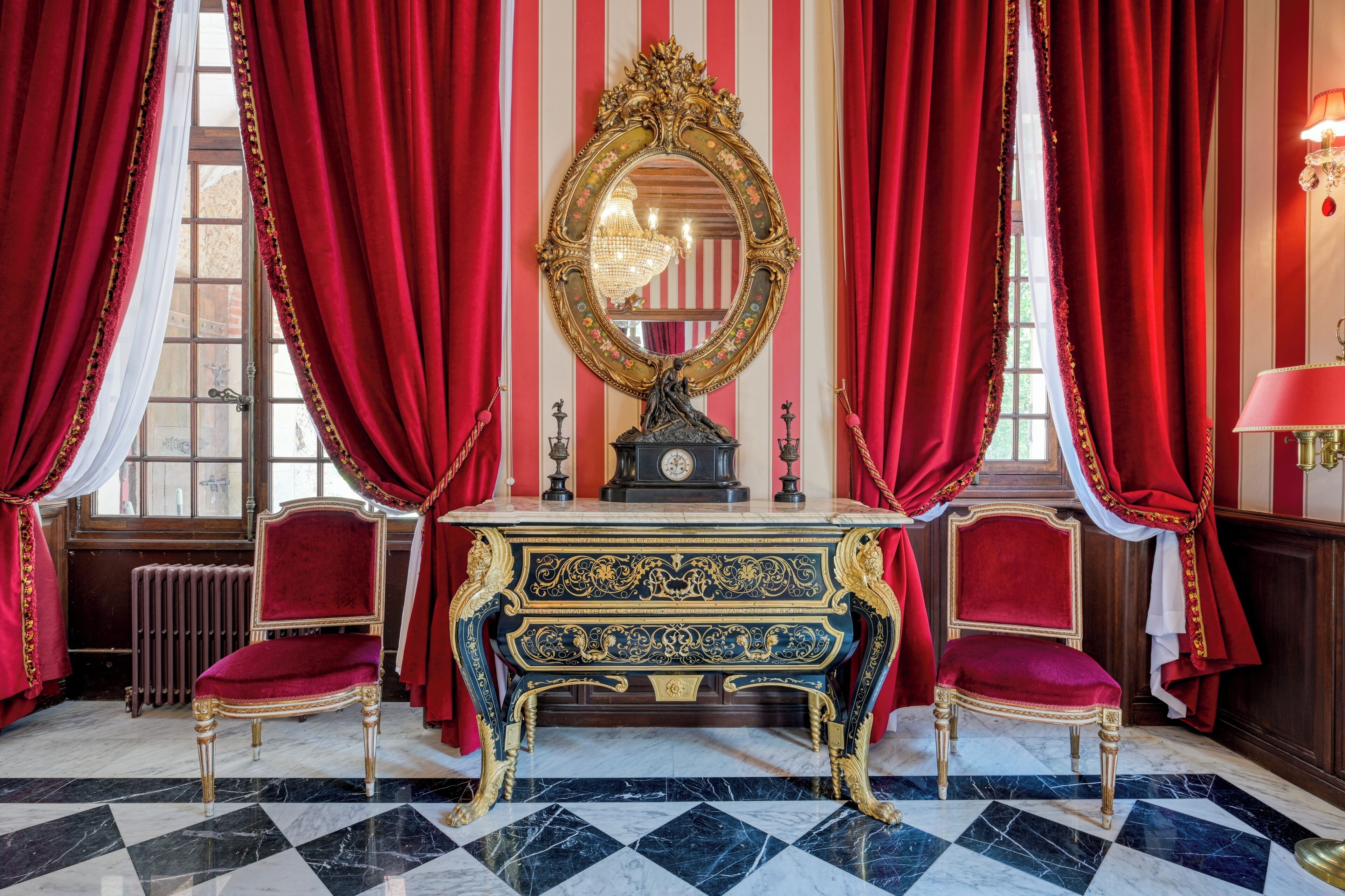 Une pièce dotée d’un grand miroir décoratif, de deux fauteuils en velours rouge, d’un sol en marbre et de rideaux rouges.