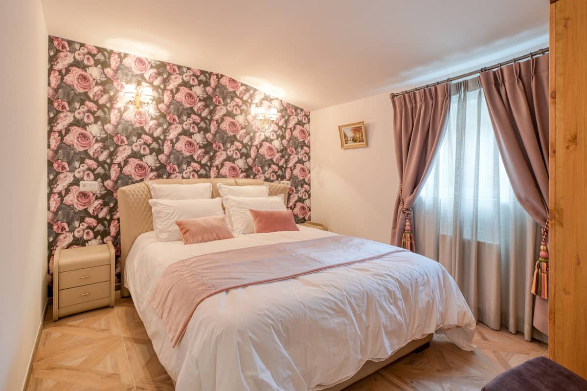Une chambre à coucher avec un papier peint à motifs floraux, un lit avec une literie blanche et rose, une table de chevet et une fenêtre avec des rideaux.