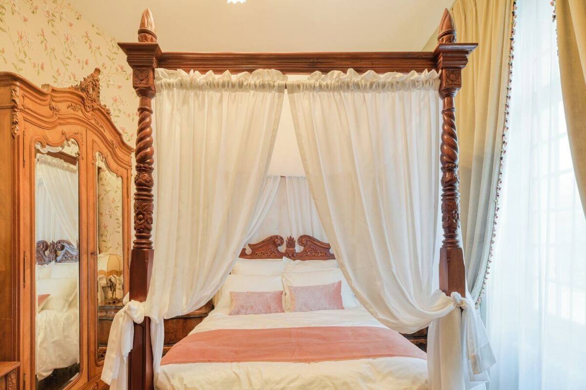 Un lit à baldaquin à quatre poteaux, avec des rideaux blancs, une tête de lit en bois et un miroir sur le côté gauche.