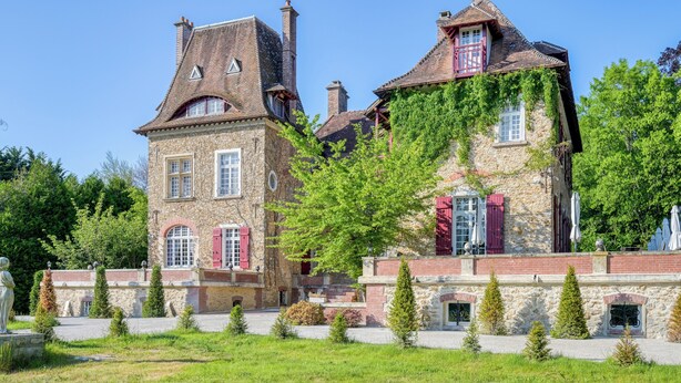 Een historisch stenen gebouw met meerdere schoorstenen, rode luiken en een goed onderhouden tuin met kleine bomen en een standbeeld.