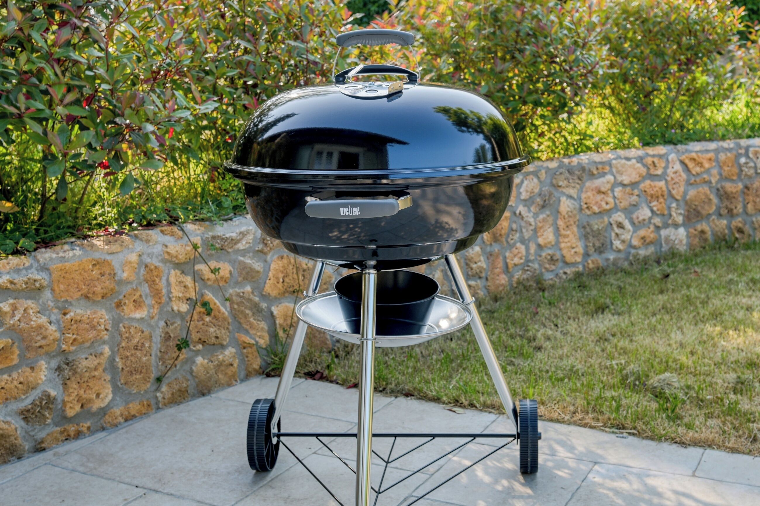 Una parrilla Weber con tapa, ubicada sobre un muro de piedra con un área verde de fondo.