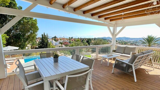 Eine Holzterrasse mit Tisch und Stühlen, mit Blick auf einen Pool und eine schöne Aussicht auf Häuser und Berge.