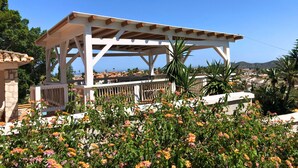 Eine weiße Pergola mit Steinunterbau, umgeben von blühenden Pflanzen und mit Blick auf ein Wohngebiet mit Häusern und Hügeln.
