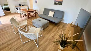 Ein modernes Wohnzimmer mit einem grauen Sofa, einem Holzesstisch, weißen Drahtstuhlen und einem kleinen Hocker. Eine Topfpflanze steht auf dem Boden und ein Gemälde hängt an der Wand.