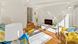En moderne stue med et fladskærms-tv, der viser 'NETFLIX', en sofa, en gul lænestol, et sofabord og en trappe, der fører til et andet rum.
