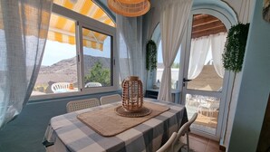 Un comedor con mantel a cuadros, sillas de plástico blanco, un farol tejido y vistas a un paisaje montañoso a través de amplios ventanales.