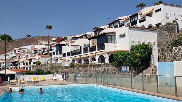 Una piscina con gente, rodeada de edificios blancos con balcones, algunos con plantas.