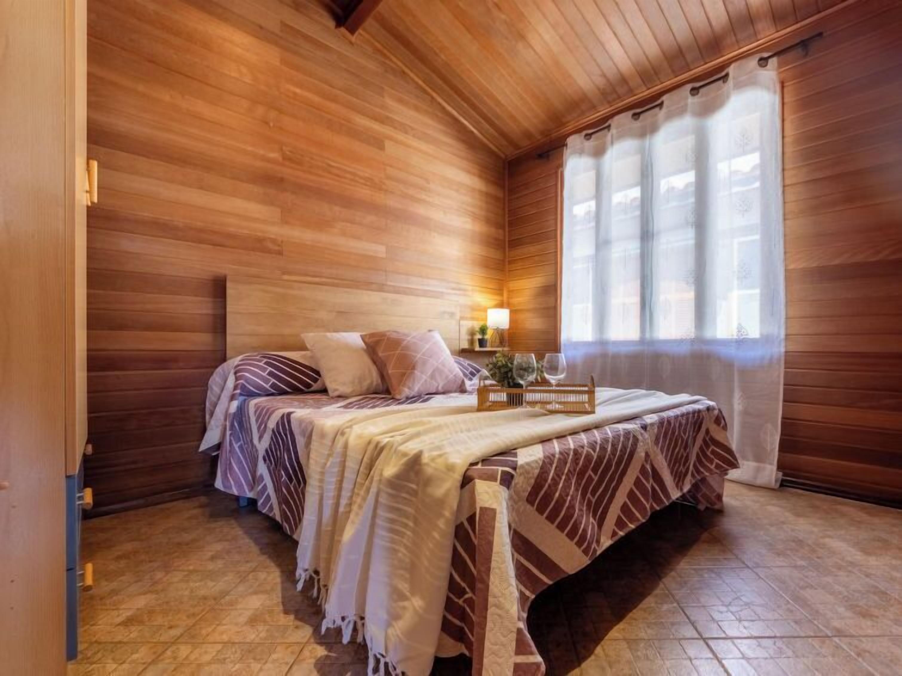 A wooden-paneled room with a bed, a bedside table, a lamp, and a window with curtains.