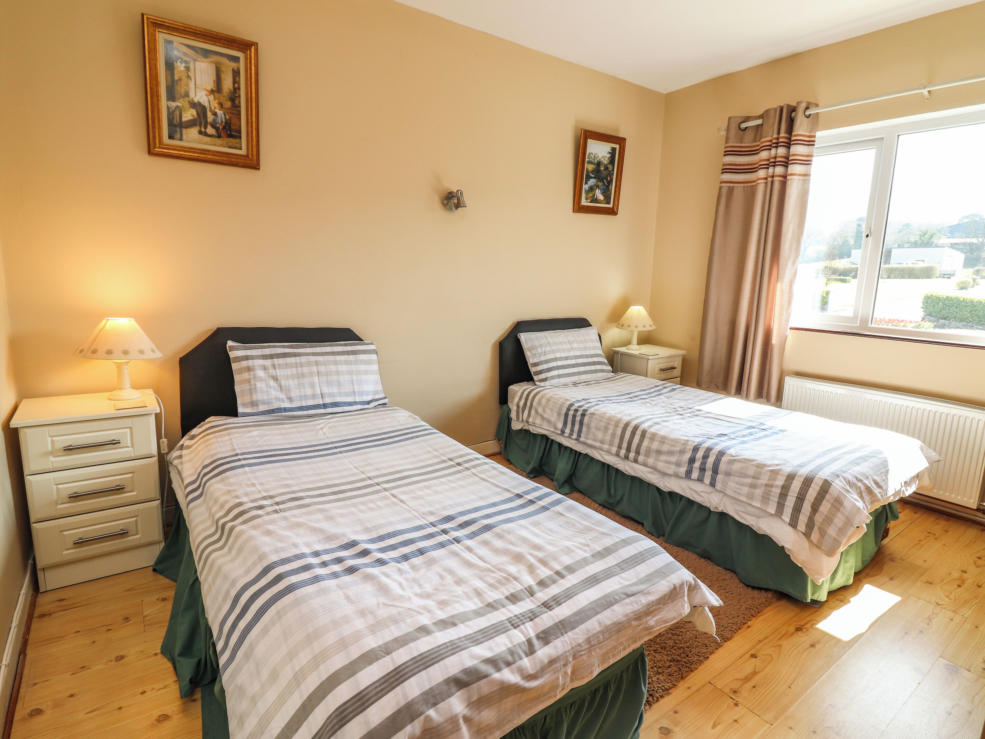A double room with two beds, each with a pillow and a bedside table with a lamp. There are two framed pictures on the wall, a window with curtains, and a radiator under the window.
