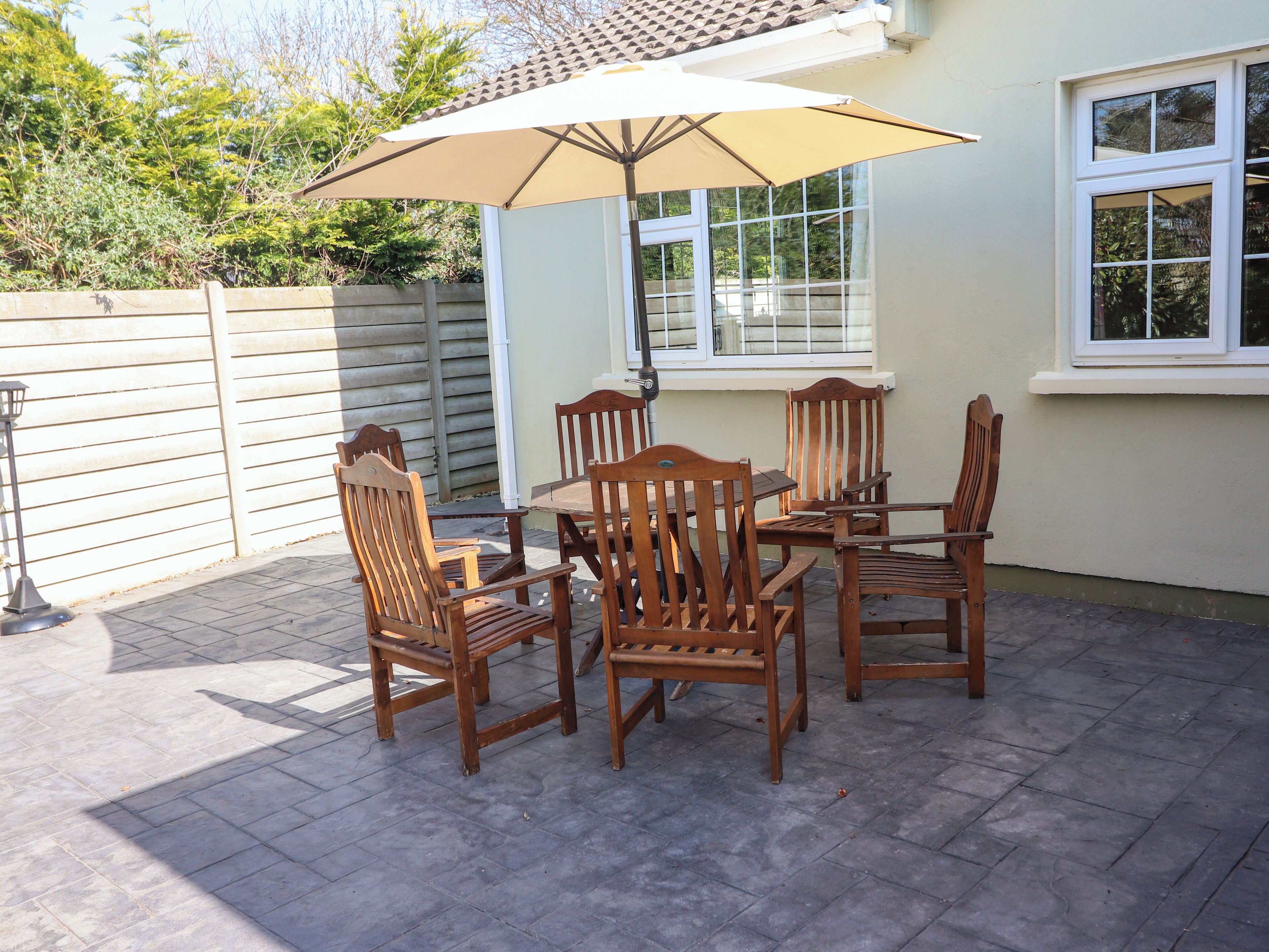 A patio with a table and chairs under a large umbrella, surrounded by a white fence and a window.