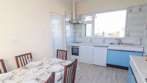 A compact kitchen with white appliances, a dining area with a table set for four, and a window overlooking the street. - Mullagh Road (Miltown Malbay)