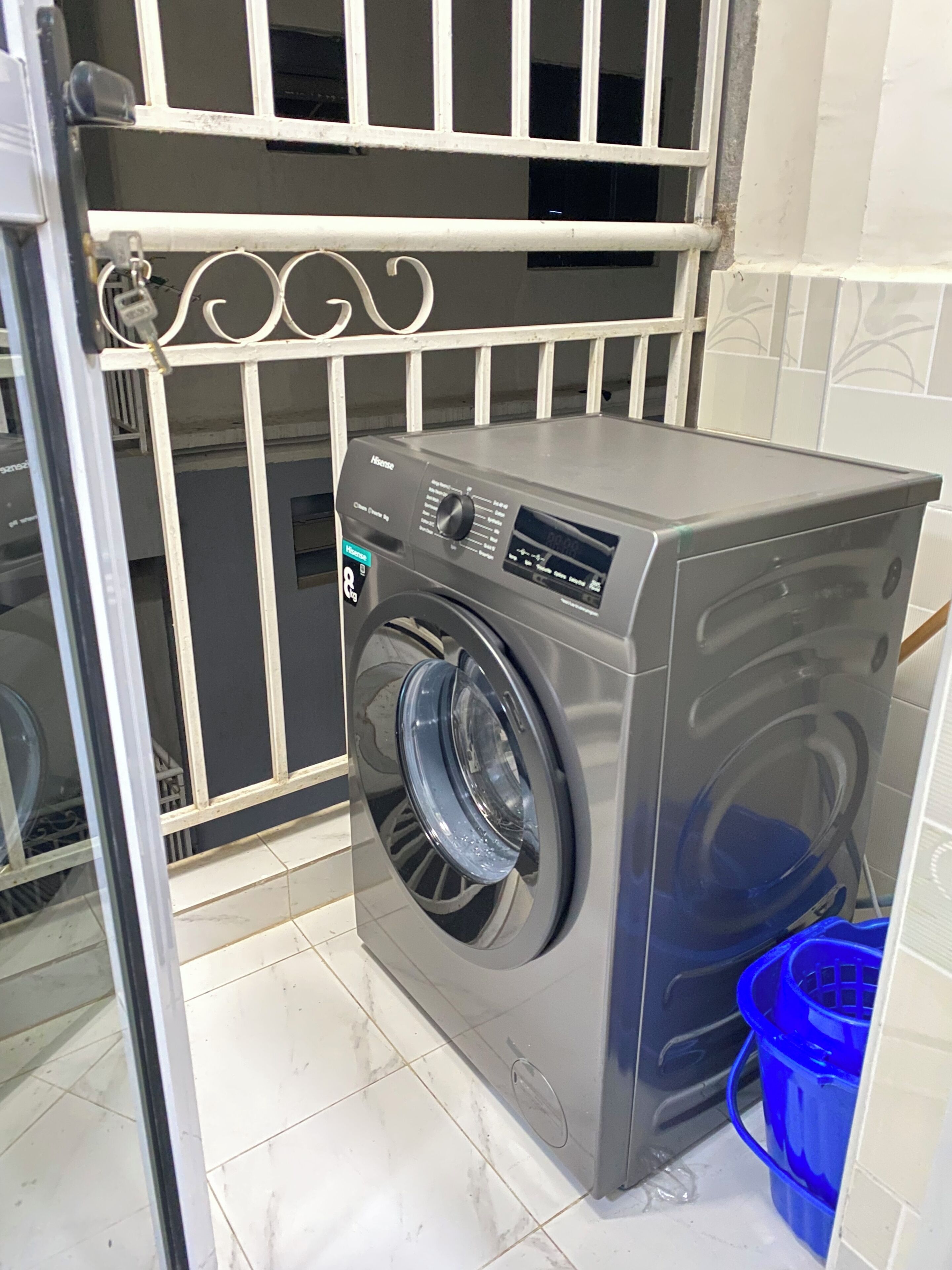 A compact, modern washing machine in a small laundry room with a metal gate and a blue plastic basket.