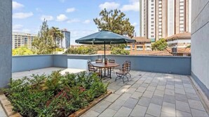 Una terraza en la azotea con mesa y sillas, rodeada de vegetación y vistas a edificios altos.