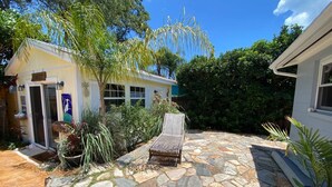 A stone patio with a lounge chair, surrounded by lush greenery and a small building with a door and windows.