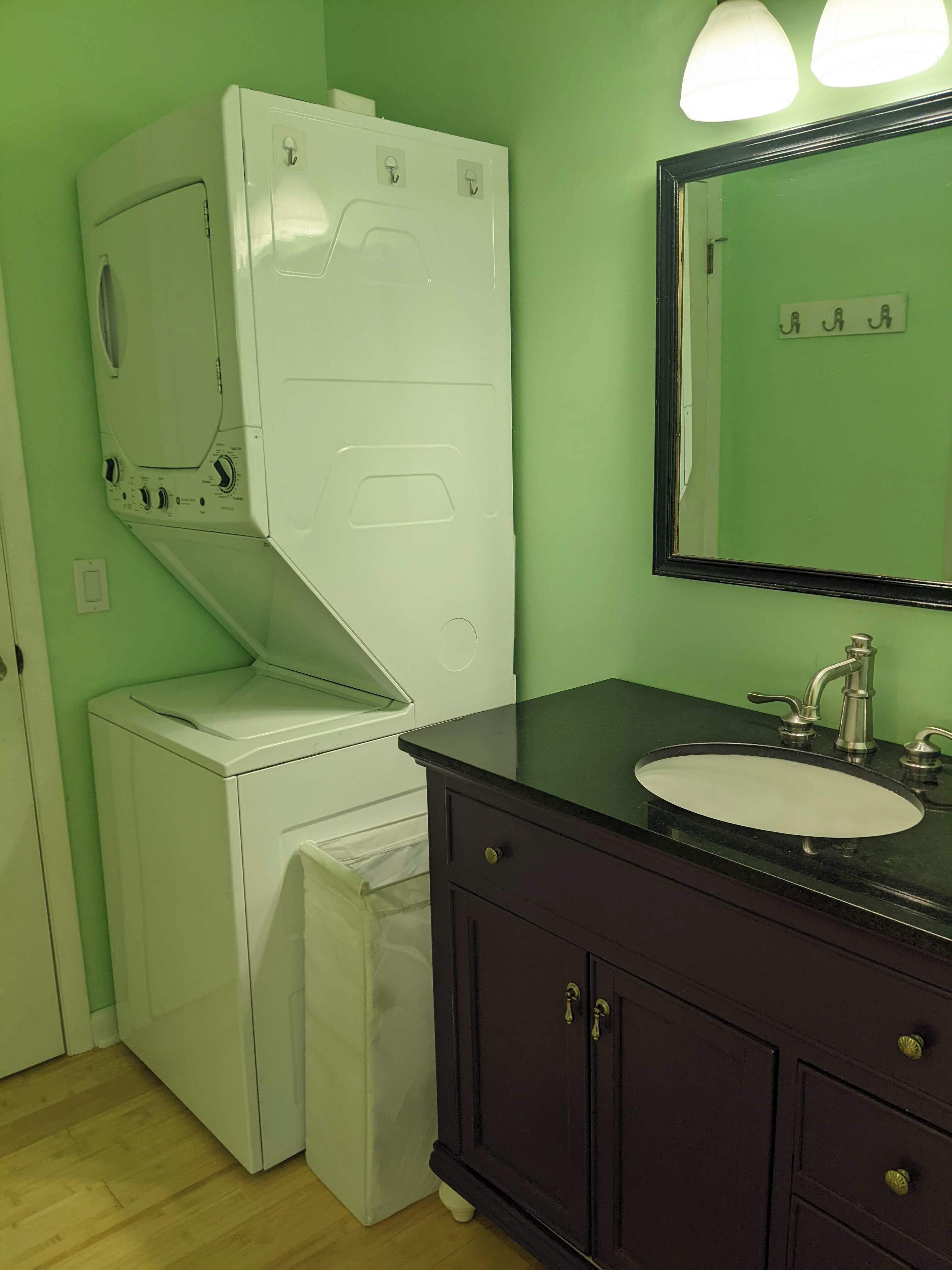 Un baño pequeño con lavadora blanca, lavamanos con encimera oscura y espejo sobre el lavamanos.