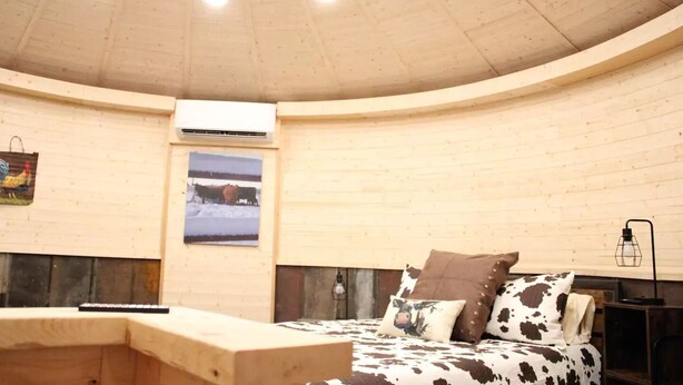 Een gezellige, ronde kamer met een houten plafond en muren, een bed met koeienprint beddengoed, een afbeelding van koeien aan de muur, en een klein tafeltje.