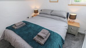 A neatly made bed with two pillows and a teal blanket, flanked by two bedside tables with lamps, a small drawer, and a framed picture on the wall.