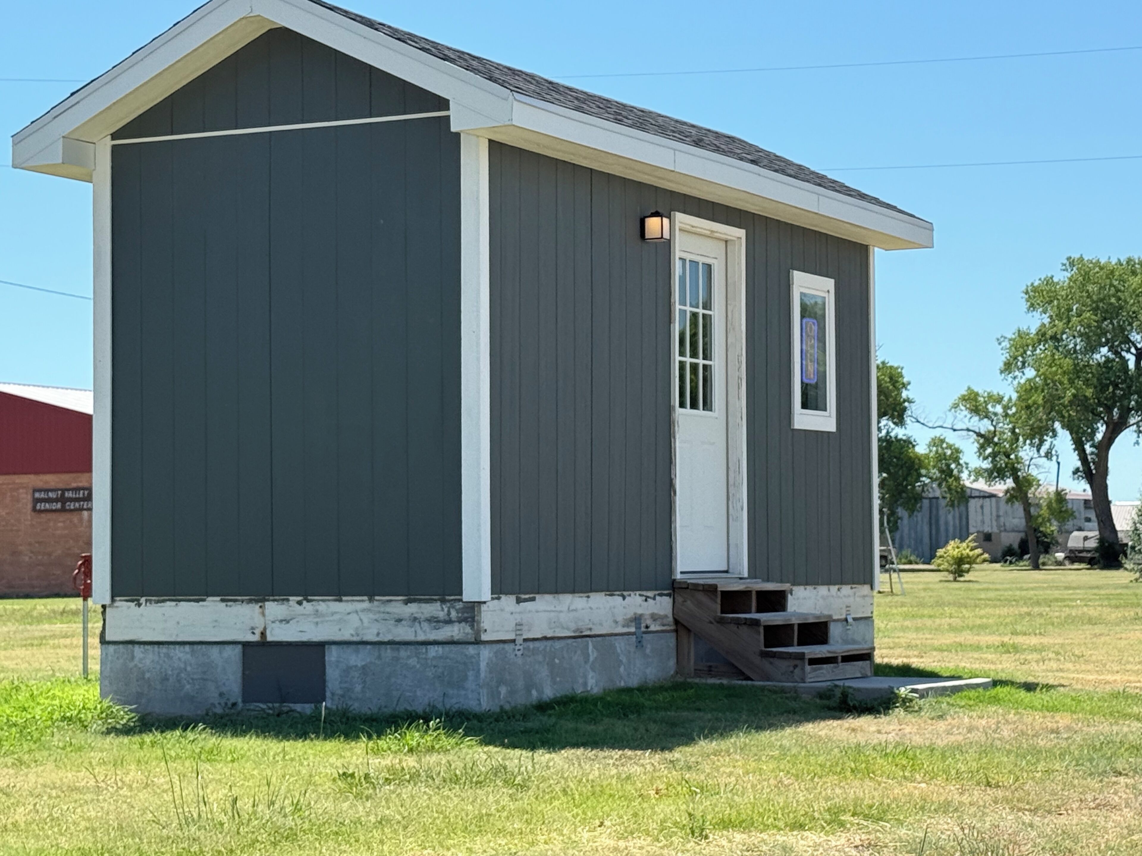 A small, single-story shed with a white door and window, situated on a grassy area with a clear sky.