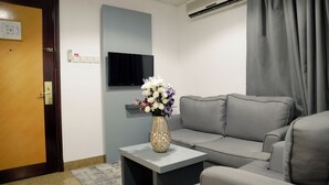 A room with a grey sofa, a small table with a vase of flowers, a flat-screen TV mounted on the wall, and an air conditioning unit. - Safeer International Hotel (Muscat)