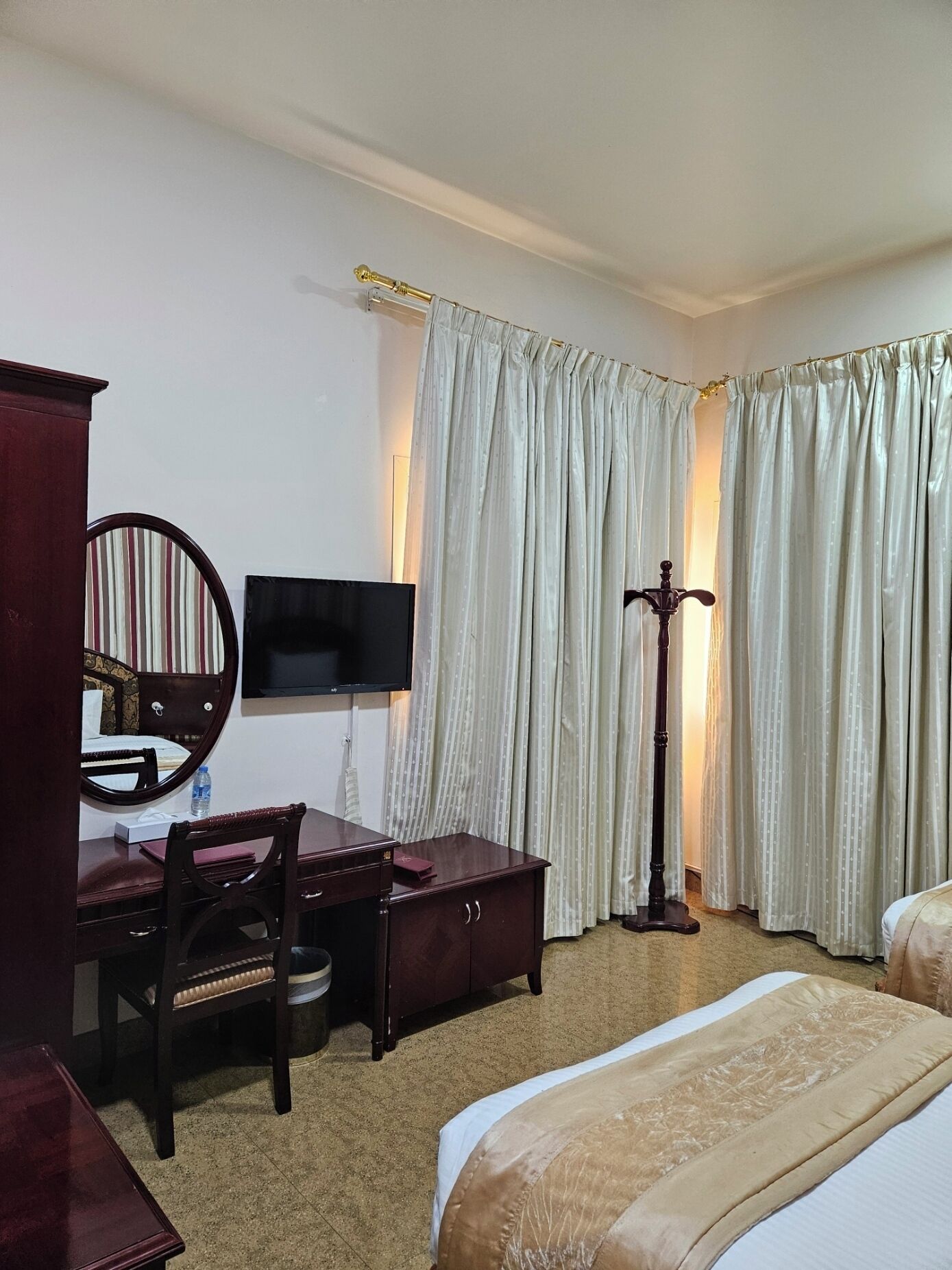 Una habitación de hotel con cama, escritorio con silla, televisor, espejo y lámpara de pie.