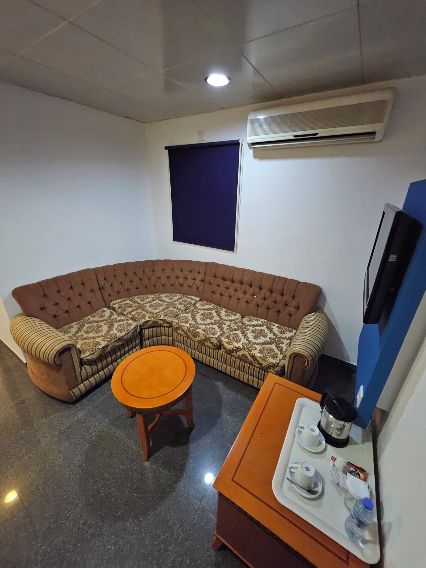 A living room with a brown sectional sofa, a round wooden coffee table, and a small side table with a tray holding a teapot, cups, and a water bottle.