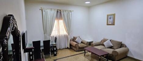 A living room with a sofa set, a dining table with chairs, a TV mounted on the wall, and a large mirror.