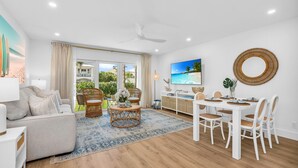 A spacious living room with a large sofa, a dining area with a white table and chairs, a ceiling fan, and a large window with a view of greenery. - ON The Beach Villa in Juno! 2! Top Rated (Juno Beach)