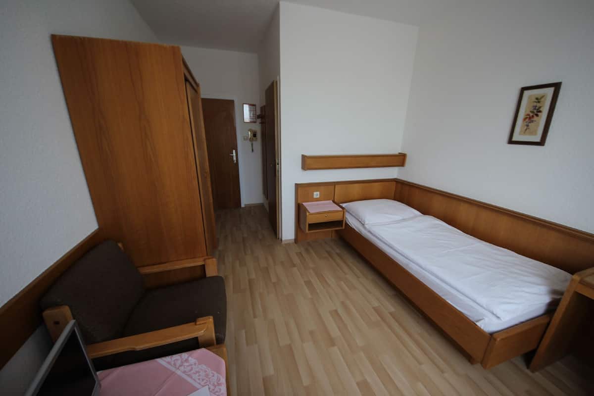 A small, compact room with a single bed, a wooden wardrobe, a small table, and a chair.
