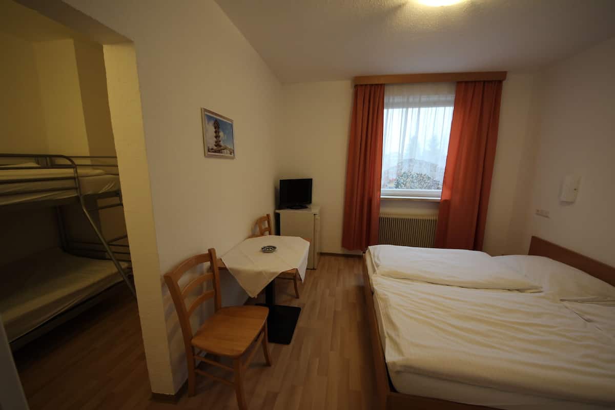 A compact hotel room with a bunk bed, a single bed, a small table with a chair, a TV, and a window with curtains.
