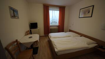 Ein kleines, ordentlich eingerichtetes Hotelzimmer mit einem Einzelbett, einem Schreibtisch mit Stuhl, einem Fernseher und einem Fenster mit Vorhängen.