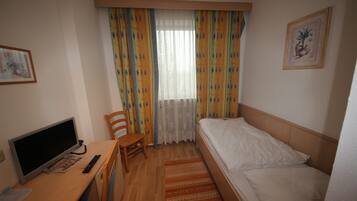 A hotel room with a single bed, a desk with a computer and remote control, a chair, and a window with curtains.