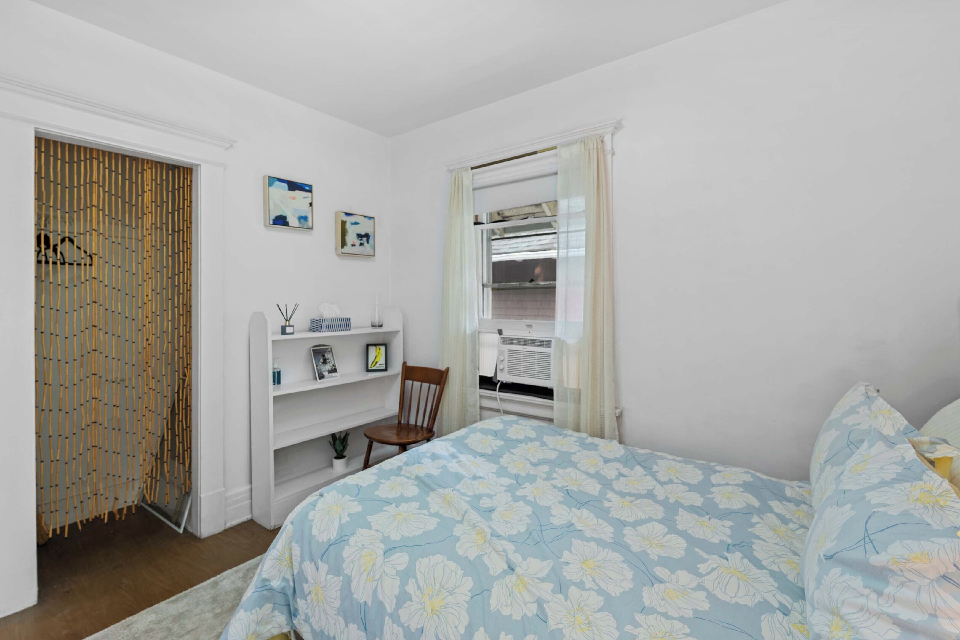 A bedroom with a bed, a chair, a shelf with items, a wardrobe, and a window with curtains.