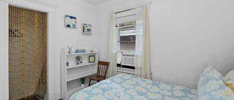 A bedroom with a bed, a chair, a shelf with items, a wardrobe, and a window with curtains.