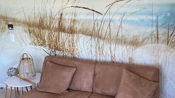 A brown sofa with cushions, a small round table with a lamp and a decorative item, and a wall mural of tall grasses.