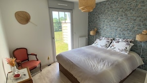 Une chambre à coucher avec un grand lit, une chaise, une petite table sur laquelle est posé un vase, et une vue sur l’extérieur.