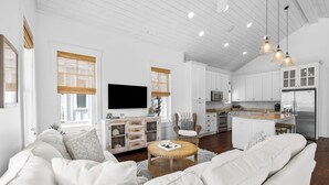 Una sala de estar moderna con un sofá, una mesa de centro y un televisor. Se aprecia una zona de cocina con armarios y un refrigerador.