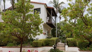 Un edificio blanco con balcón, rodeado de abundante follaje y plantas florecientes.